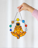 Hand holding a stained glass suncatcher of an astronaut with colorful border against a white curtain.