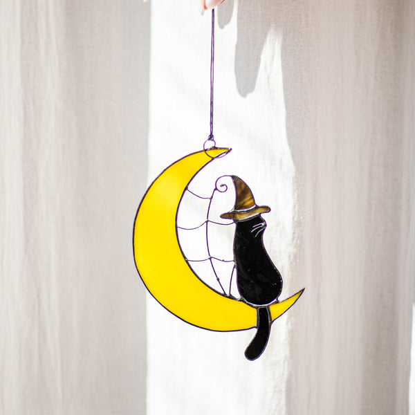 Decorative hanging with a black cat in a witch hat on a yellow moon against a white curtain.