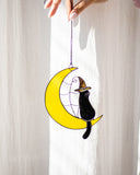 Decorative hanging with a black cat in a witch hat on a yellow moon against a white curtain.