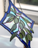A stained glass suncatcher featuring a blue border and green olive branches with iridescent white and blue reflective panels, hanging in front of a window.