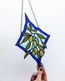 A stained glass suncatcher featuring an olive branch design with green olives on a light blue and dark blue background, held by hands against a white backdrop.