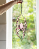 A hand holds a pink spider suncatcher with black metal and glass detailing in front of a bright window with a blurred green outdoor background.