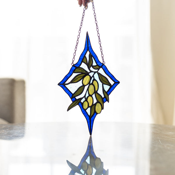 A stained glass suncatcher featuring an olive branch design with blue borders, green leaves, and yellow-green olives, held by a hand over a reflective glass table.