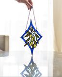 A stained glass suncatcher featuring an olive branch design with blue borders, green leaves, and yellow-green olives, held by a hand over a reflective glass table.
