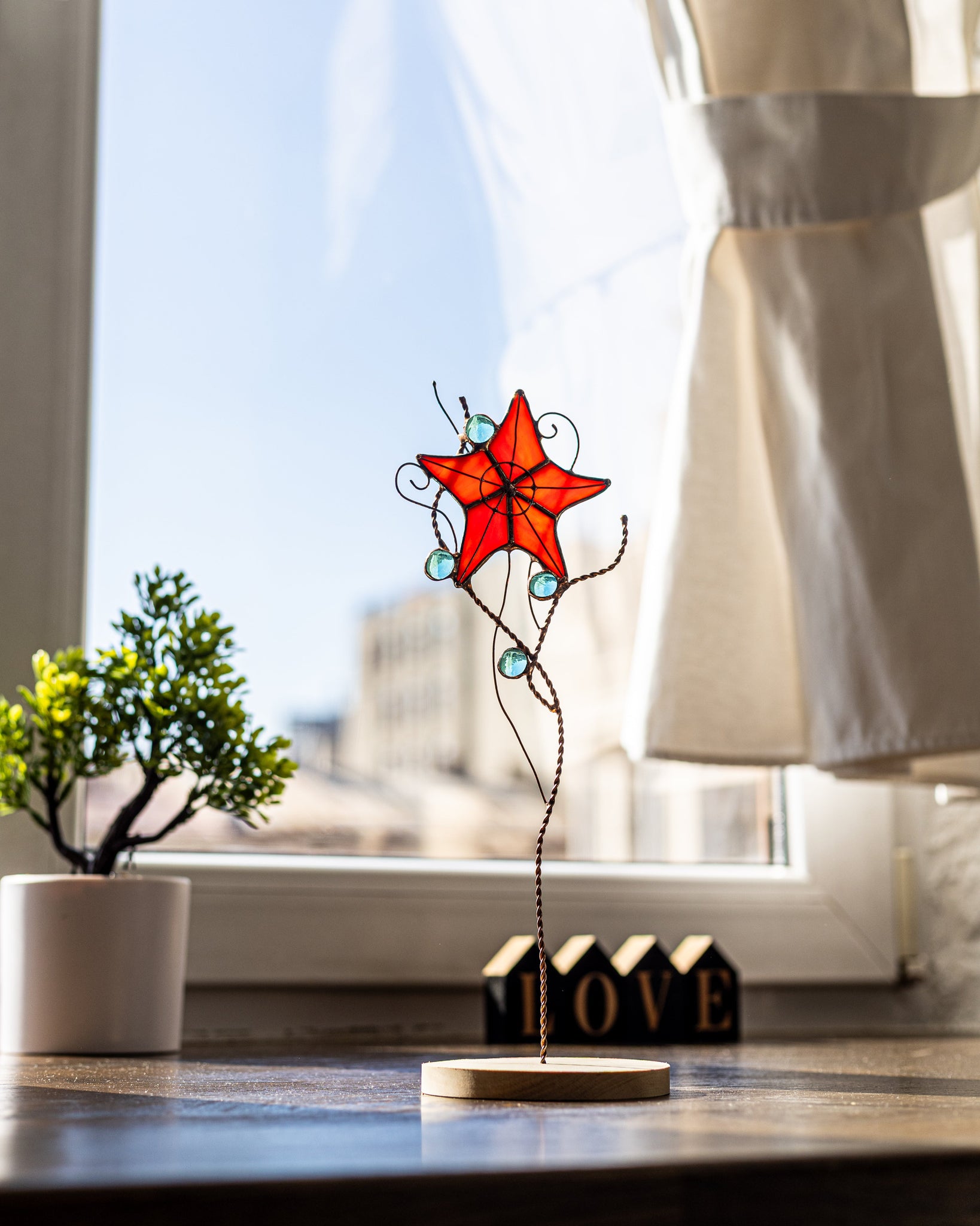 Handmade stained glass star flower on copper wire stem with blue glass gems, displayed in natural sunlight on a wooden base by the window — unique Tiffany-style decor for plant pots, vases, garden beds, or as whimsical boho home art.