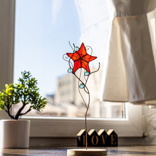 Handmade stained glass star flower on copper wire stem with blue glass gems, displayed in natural sunlight on a wooden base by the window — unique Tiffany-style decor for plant pots, vases, garden beds, or as whimsical boho home art.