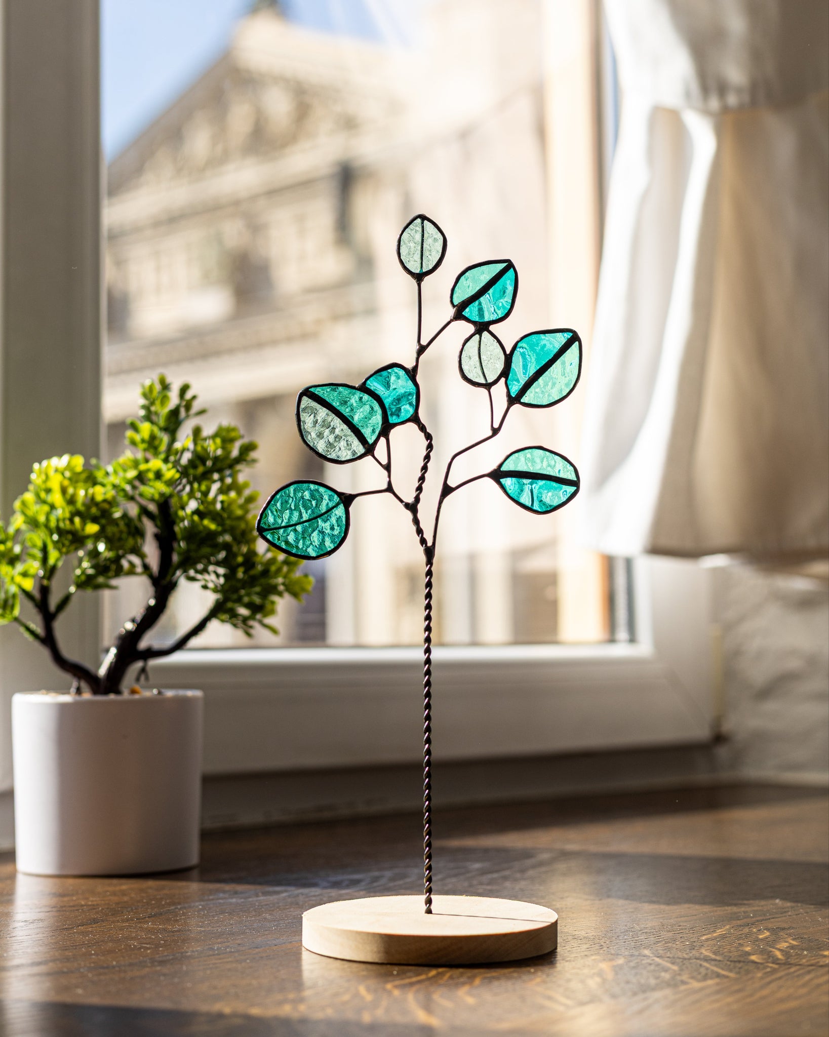 Decorative glass plant with teal leaves on a wooden base in a room with a window.