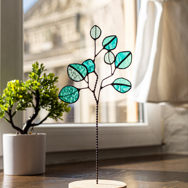 Decorative glass plant with teal leaves on a wooden base in a room with a window.