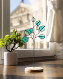 Decorative glass plant with teal leaves on a wooden base in a room with a window.
