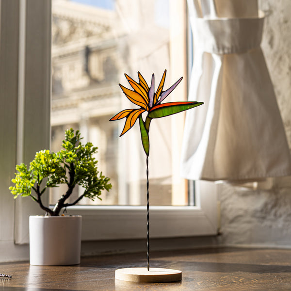 Decorative stained glass bird of paradise flower on a wooden base in front of a window with curtains.
