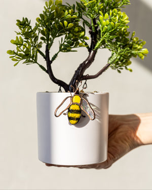 Hand holding a white pot with a small plant and a bee-shaped keychain on a light background