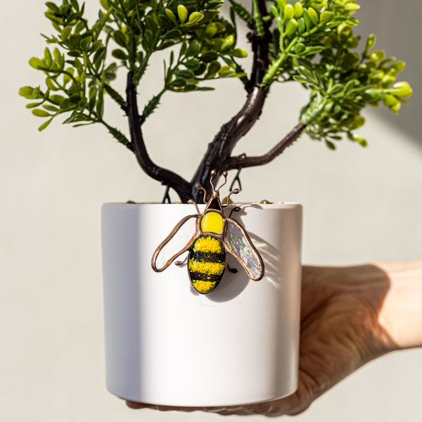 Hand holding a white pot with a small plant and a bee-shaped keychain on a light background
