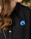 A close-up of a woman wearing a black collared shirt or jacket with a bright blue flower-shaped brooch pinned near the chest.