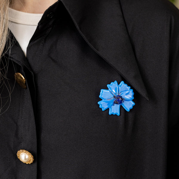 A close-up of a person wearing a black collared shirt or jacket with gold buttons and a bright blue flower-shaped brooch pinned near the chest.