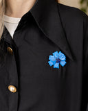 A close-up of a person wearing a black collared shirt or jacket with gold buttons and a bright blue flower-shaped brooch pinned near the chest.