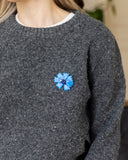 A close-up of a woman wearing a dark gray knit sweater with a blue flower-shaped brooch pinned to the chest, set against a softly blurred indoor background.