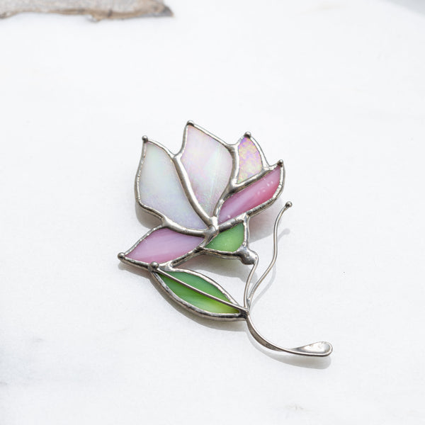 A stained glass flower brooch with pink, white, and green enamel panels and silver metal borders, resting on a white marble surface.