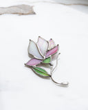 A stained glass flower brooch with pink, white, and green enamel panels and silver metal borders, resting on a white marble surface.