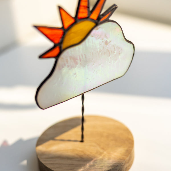 A stained-glass-style sun and cloud ornament on a round wooden base, photographed on a bright white surface with soft natural light.
