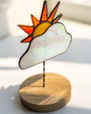 A stained-glass-style sun and cloud ornament on a round wooden base, photographed on a bright white surface with soft natural light.