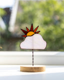 A stained glass sun and cloud suncatcher on a twisted metal stand with a round wooden base, displayed on a windowsill with a blurred outdoor garden background.
