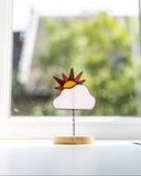 A small decorative stained-glass-style sun and cloud ornament on a thin stand with a round wooden base, placed on a windowsill with a blurred outdoor scene behind it.