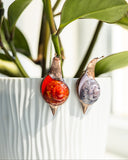 Two decorative snail ornaments with colorful shells hang from green plant stems in a white ribbed planter.