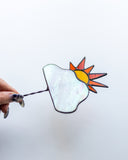 A stained glass suncatcher featuring a white mother-of-pearl cloud and a yellow and orange sun, held by a hand against a plain white background.