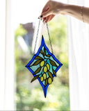 A hand holds a stained glass suncatcher featuring a blue diamond frame, green leaves, and yellow-green olives, positioned in front of a bright window.