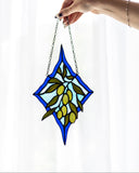 A hand holds a stained glass suncatcher featuring an olive branch design with green leaves and yellow-green olives, set against a light blue and dark blue diamond-shaped background with a silver chain.