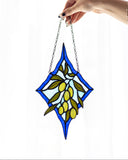 A hand holds a stained glass suncatcher featuring an olive branch design with green leaves and yellow-green olives, set against a blue and white diamond-shaped border with a silver hanging chain.