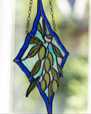 A stained glass suncatcher featuring a green olive branch design with a blue diamond border, hanging from a metal chain in front of a softly blurred window background.