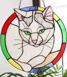 Stained glass cat design hanging on a white wall with plants in the foreground