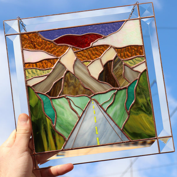 Stained glass panel with mountain and road design held against a blue sky.