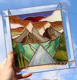 Stained glass panel with mountain and road design held against a blue sky.