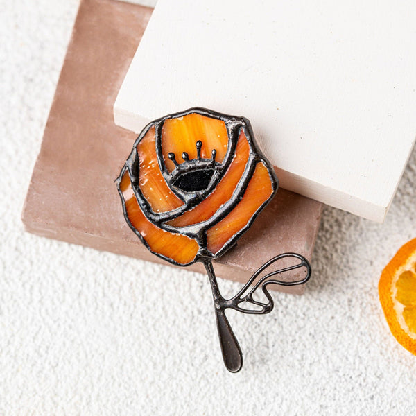 An orange rose-shaped brooch with black metal outlines and a small crown detail, displayed on a brown and white square card with dried orange slices on a textured white surface.