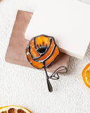 An orange rose-shaped brooch with black metal outlines and a small crown detail, displayed on a brown and white square card with dried orange slices on a textured white surface.