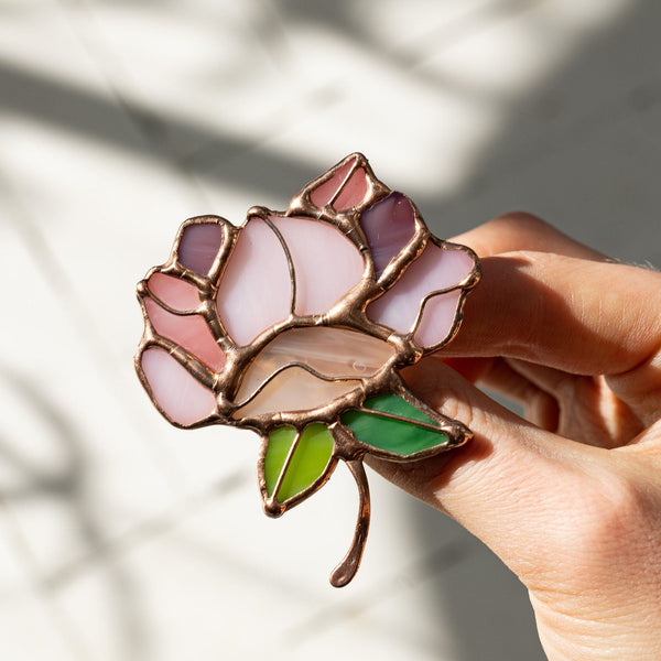 A hand holding a stained glass rose brooch with pink, purple, and green glass panels outlined in gold-toned metal, set against a softly shadowed light background.
