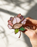 A hand holding a stained glass rose brooch with pink, purple, and green glass panels outlined in gold-toned metal, set against a softly shadowed light background.