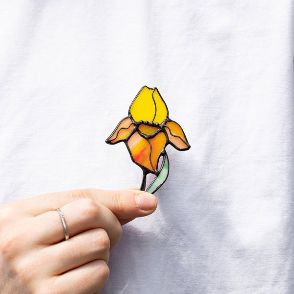 A hand wearing a silver ring holds a small embroidered flower pin against a white shirt. The flower has yellow, orange, and peach petals with a green stem and black outlines.