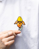 A hand wearing a silver ring holds a small embroidered flower pin against a white shirt. The flower has yellow, orange, and peach petals with a green stem and black outlines.