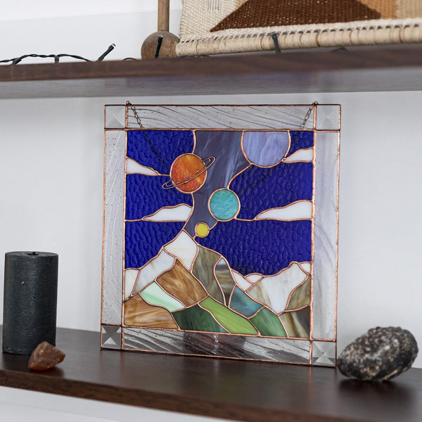 Stained glass panel of a landscape with mountains and celestial bodies on a shelf.