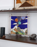 Stained glass panel of a landscape with mountains and celestial bodies on a shelf.