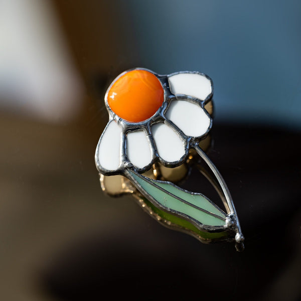 A close-up of a flower-shaped enamel pin with white petals, a bright orange center, and green leaves outlined in silver metal, resting on a dark reflective surface.