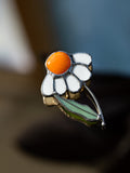 A close-up of a flower-shaped enamel pin with white petals, a bright orange center, and green leaves outlined in silver metal, resting on a dark reflective surface.