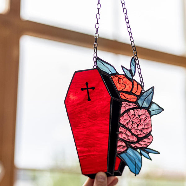 A coffin-shaped stained glass window panel with a black cross, red glass, pink brain roses, and blue leaves, suspended by a chain and held by hands in front of a softly blurred window.