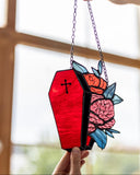 A coffin-shaped stained glass window panel with a black cross, red glass, pink brain roses, and blue leaves, suspended by a chain and held by hands in front of a softly blurred window.
