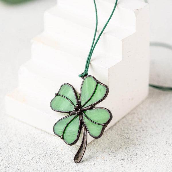 A green four-leaf clover pendant with dark metal outlines, hanging from a green cord, displayed on a white geometric block against a light gray textured background.
