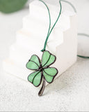 A green four-leaf clover pendant with dark metal outlines, hanging from a green cord, displayed on a white geometric block against a light gray textured background.