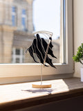 A black and silver abstract metal wall art sculpture with a wooden base, displayed on a wooden windowsill in bright natural light.